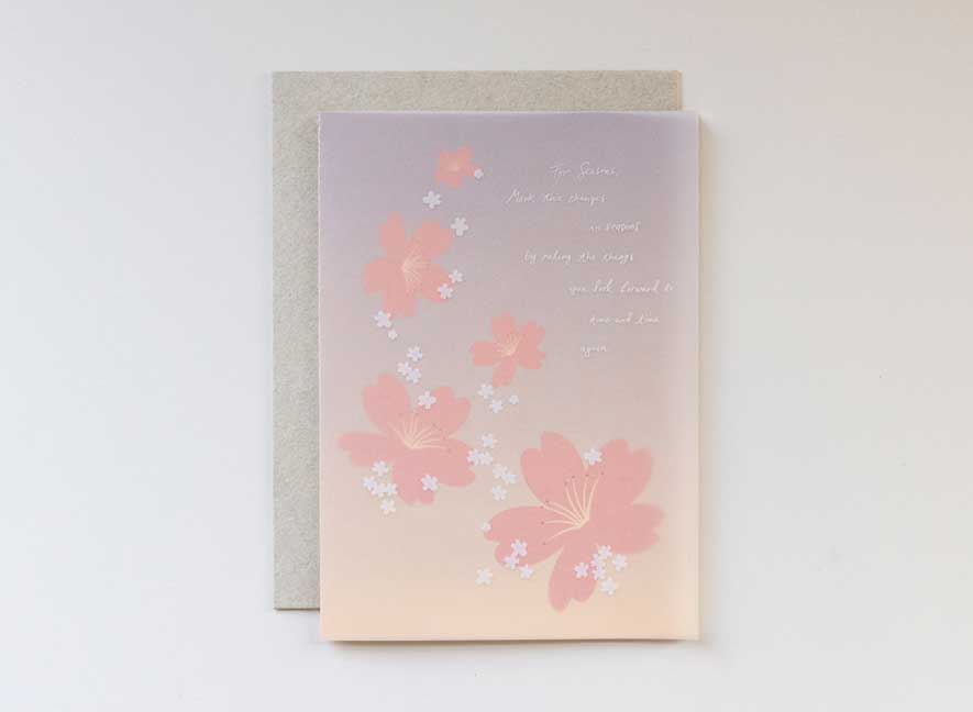 Luxury greeting card. Two-ply card with a translucent light pink cover printed with small white cherry blossoms that cascade throughout the paper. Interior is cream cardstock printed with blue skies larger and large white cherry blossoms that have been pressed into the paper. English-only version.