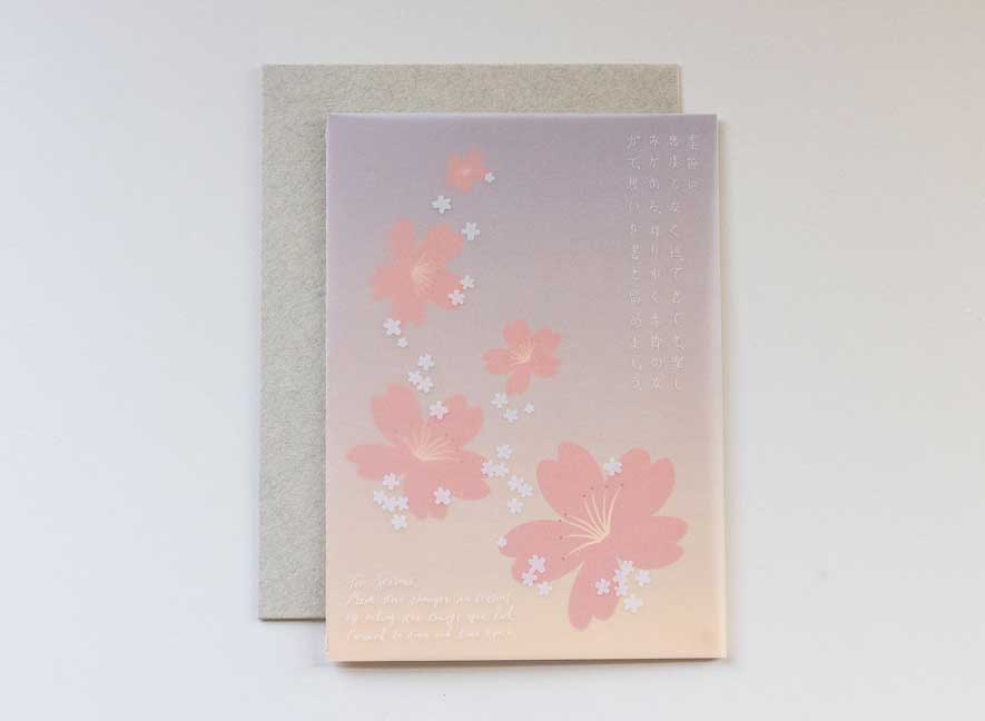 Luxury greeting card. Two-ply card with a translucent light pink cover printed with small white cherry blossoms that cascade throughout the paper. Interior is cream cardstock printed with blue skies larger and large white cherry blossoms that have been pressed into the paper. English-Japanese version.